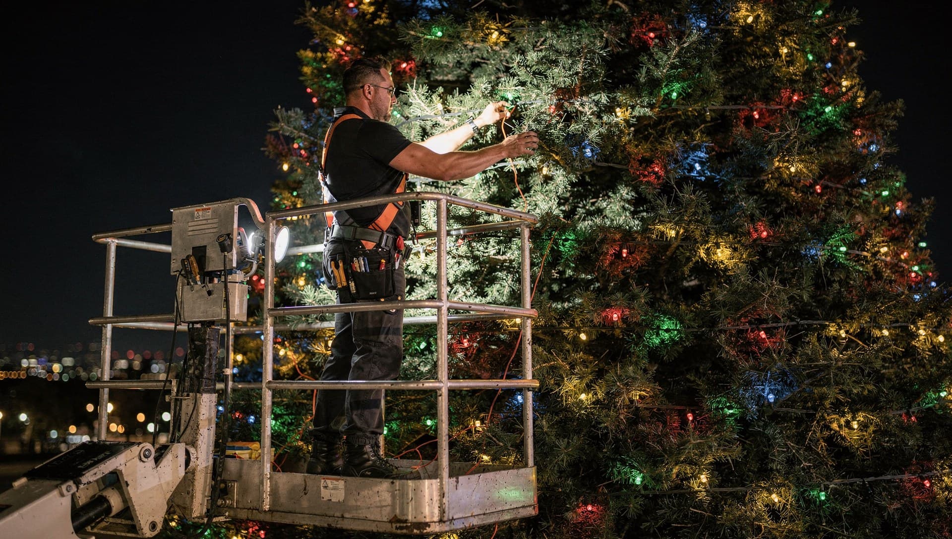 Professional installer on bucket lift installing colorful Christmas lights on illuminated tree at night